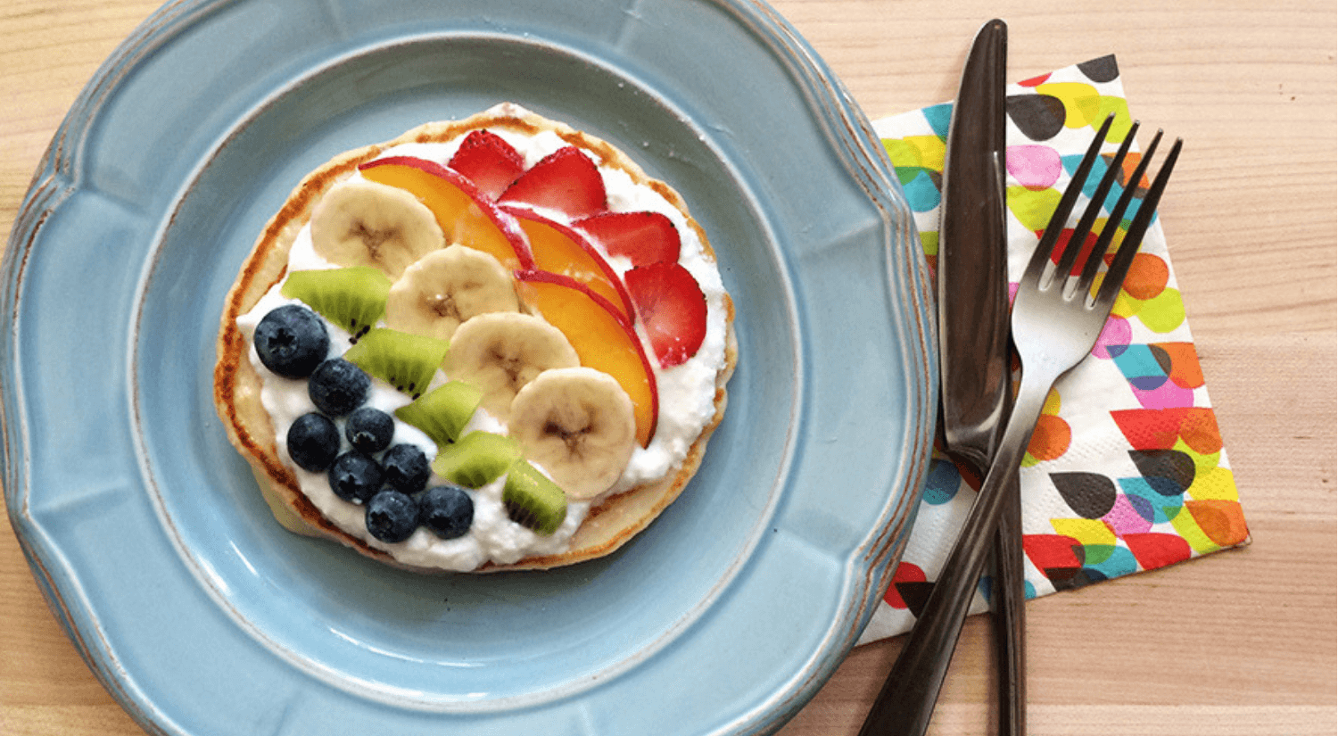 Blue plate with a pancake topped with greek yogurt, blueberries, kiwi, bananas, peaches and strawberries to make a fruit rainbow.