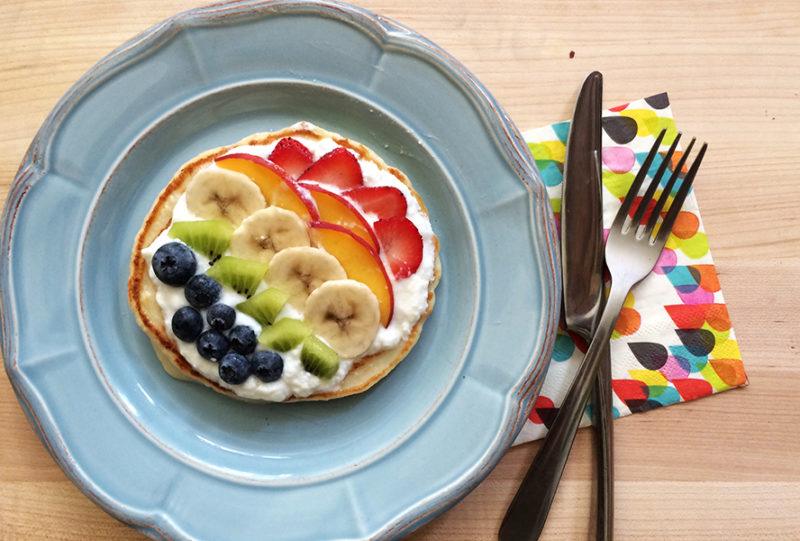 Blue plate with a pancake topped with greek yogurt, blueberries, kiwi, bananas, peaches and strawberries to make a fruit rainbow.